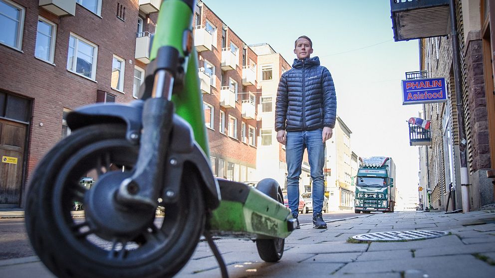Johan Larsson – en man i jeans och dunjacka – står bakom en elsparkcykel parkerad mitt på en trottoar.