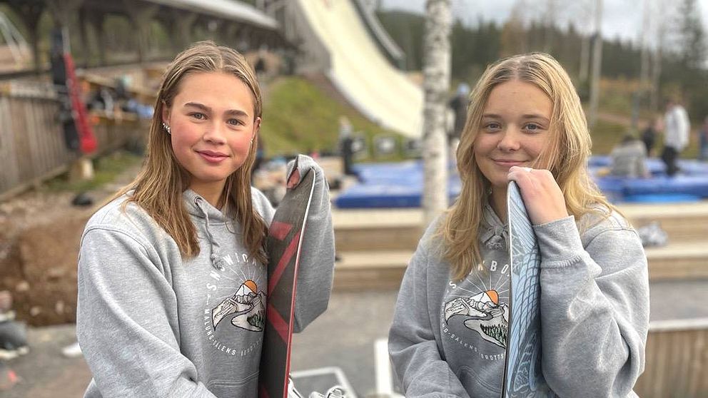 Två tonårstjejer som står och hänger på var sin snowboard utomhus, det är barmark och i bakgrunden skymtar en konstgjord skidbacke.