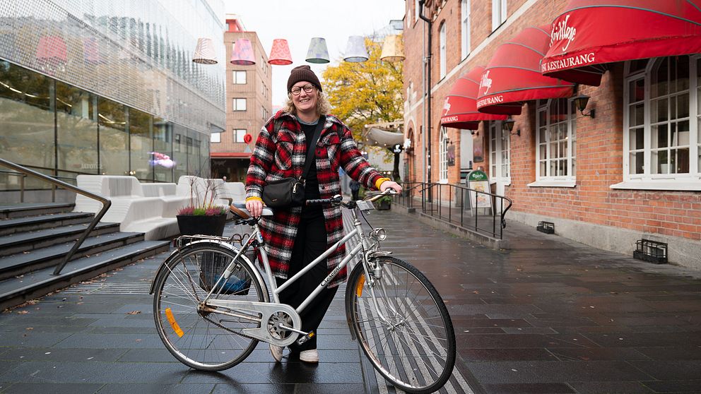 Helbild på kvinna, blont hår, brun mössa, glasögon, kappa i i rött och vitt, står och håller sin cykel på en gågata med grå stenplattor.