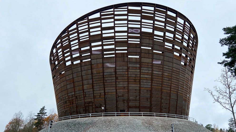 En modern, rund byggnad med ett öppet träverk som liknar en stor korg, belägen på en kulle omgiven av träd och naturlandskap. Byggnaden har en unik design med stora öppningar som släpper in ljus.