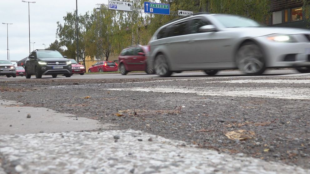 En asfalterad väg med mycket trafik, några av bilarna swishar suddigt förbi. I bakgrunden syns vägskyltar mot centrum, E4 södra och Backen.