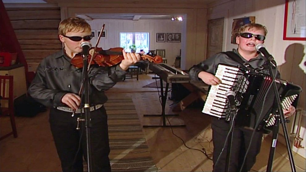Två barn i matchande grå silkesskjortor och solglasögon spelandes på fiol och dragspel