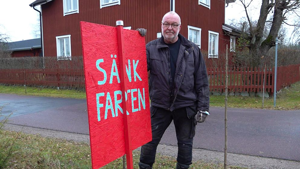 Bild på Olle Eriksson som ser bestämd ut när han står bredvid vägen och håller handen på en stor röd skylt med texten ”SÄNK FARTEN” skriven i vit text med versaler.
