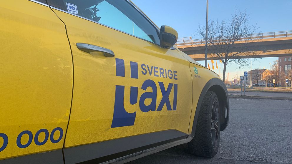 På bilden syns en taxibil från företaget Sverigetaxi. Himlen är blå och till höger i bilden i bakgrunden syns en bro.