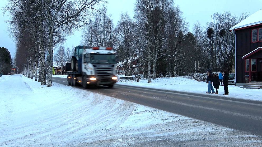 En lastbil lastad med en traktor kör på en landsväg. Några barn väntar på att kunna passera.