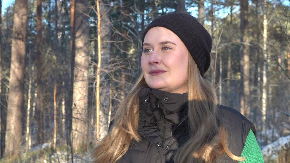 Bild på kvinna med svart mössa som står i en skog. Kvinnan heter Maja Johansson och ska i sitt examensarbete undersöka hur Storforsborna ser på skogen.