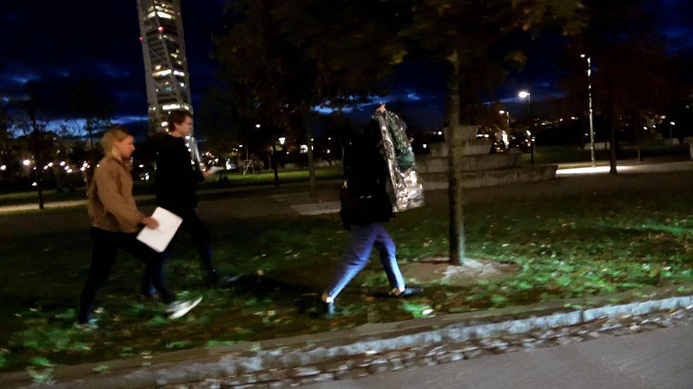 Två av SVT:s reportar följer efter Soheil Naderi i en park i Malmö på kvällen. Turning torso syns i bakgrunden.