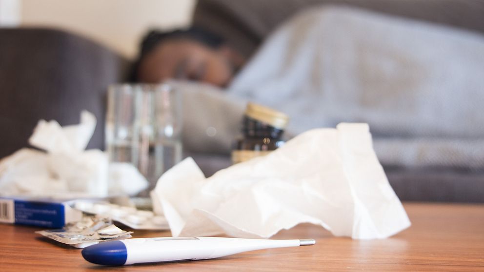 En anonym man ligger sjuk på soffan med medicin, snytpapper och termometer framför sig på bordet.