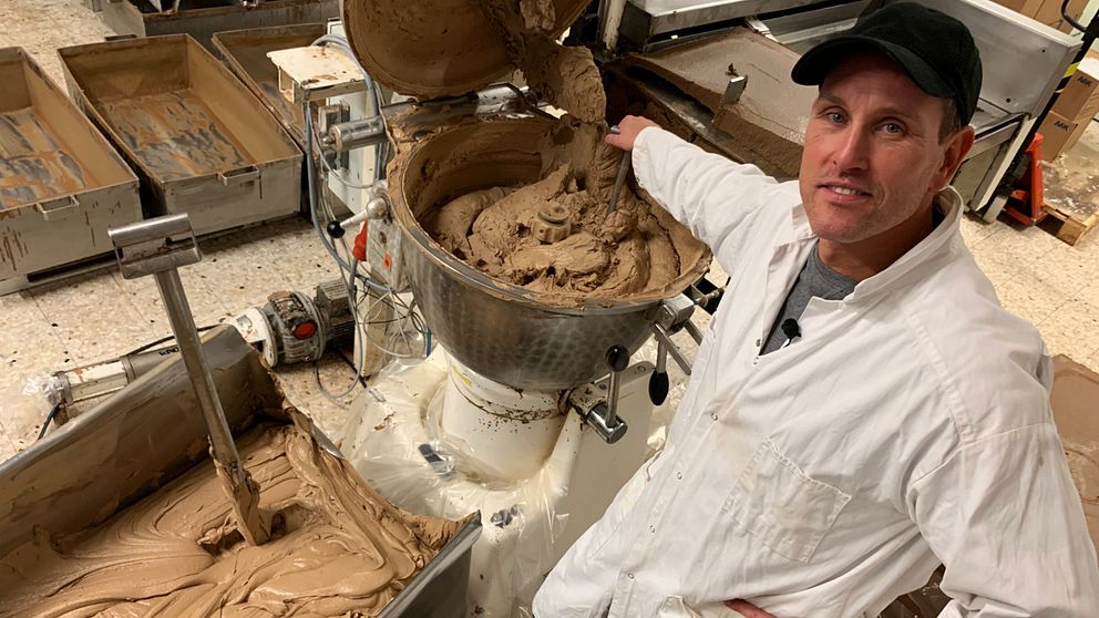 Bild på en man i vit rock som står vid en maskin med brun smet. Mannen jobbar på nötkrämsfabriken i Hässleholm där tonvis med hasselnöter slinker ner i maskinen varje månad.