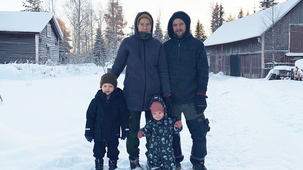 En mamma, pappa, son och dotter. I bakgrunden ser man en ladugård och ett gammalt timmerhus. Det ser kallt ut och det är mycket snö på marken.