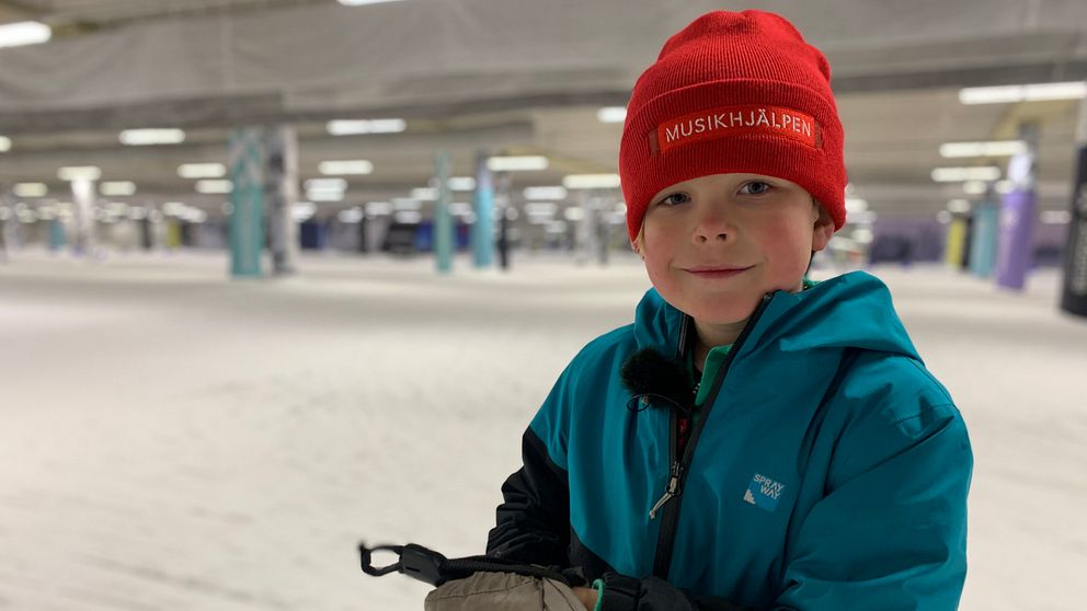 Bild på en pojke med röd mössa där det står Musikhjälpen. Pojken heter Arvid och står i en anläggning för längdskidor där han ska åka för att samla ihop pengar.