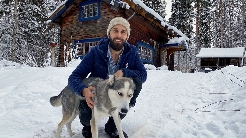 Youtubern Kalle Flodin sitter på huk i snön och håller sin huskey Tuss i famnen. I bakgrunden ser vi hans timmerstuga.