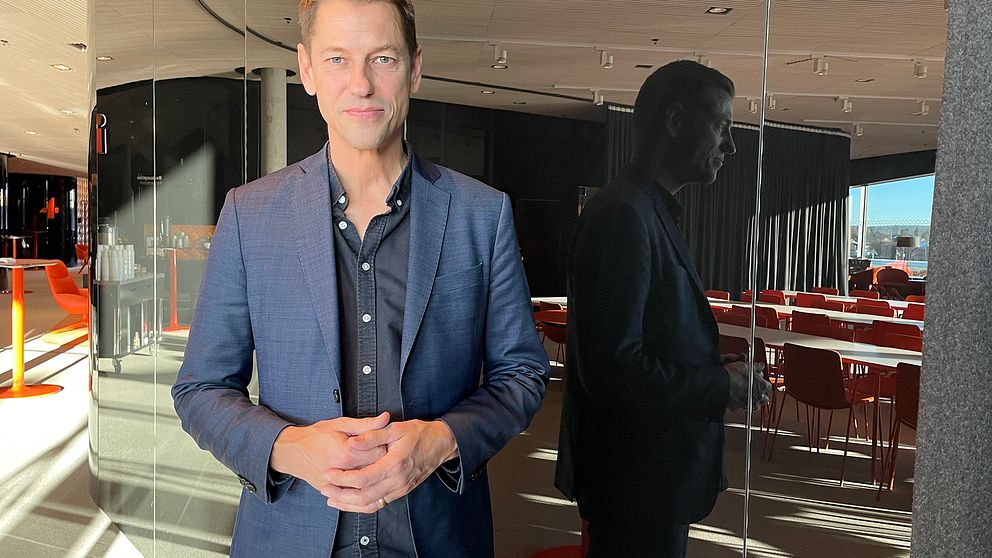 En man står med händerna knäppta framför magen. Bakom honom reflekteras hans profil i en spegelvägg.