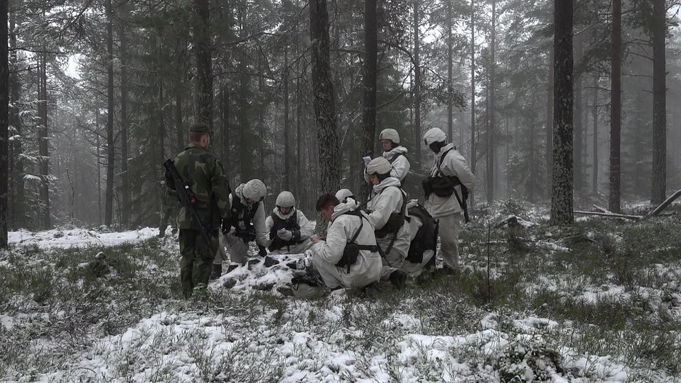 Soldater i vinterkamouflage i skog, diskuterar karta och strategi.