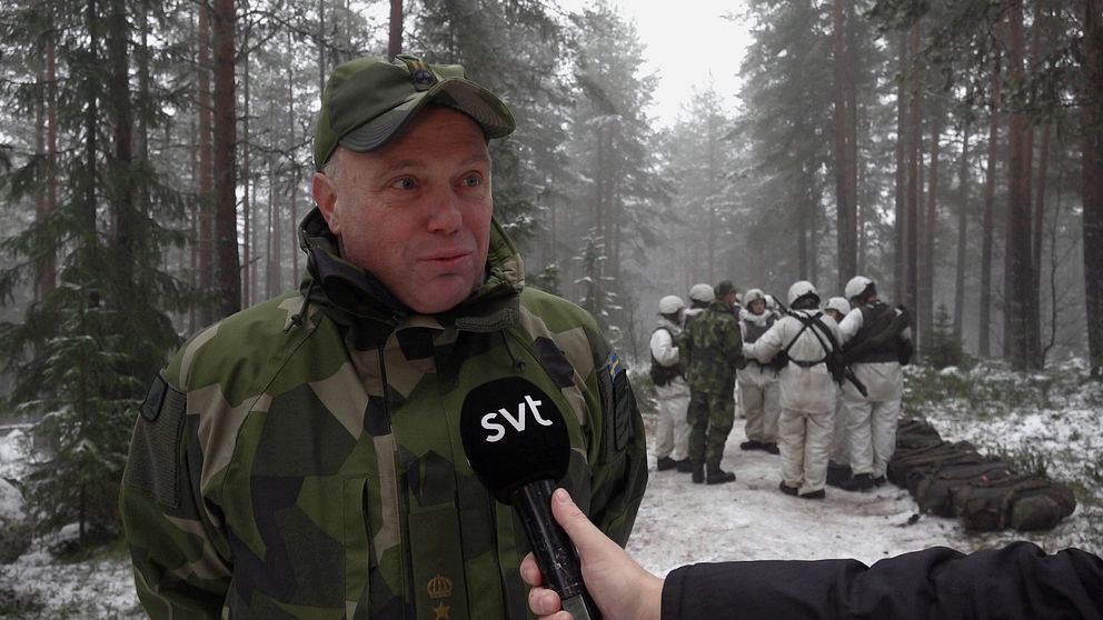 Reporter intervjuar soldat i vinterkamouflage i skogsmiljö, fler soldater i bakgrunden.