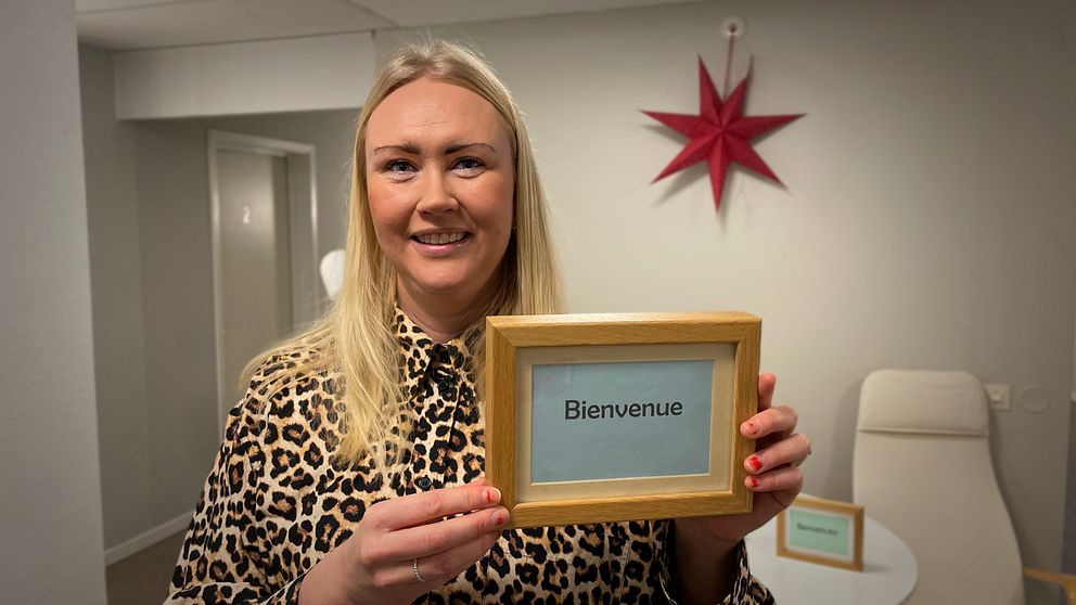 Bild på en blond kvinna med en skylt där det står Bienvenue. Kvinnan heter Elina Perdsjö och är verksamhetsledare för natthärbärget Vinternatt. Hon bjuder på en rundtur i de nya lokalerna.