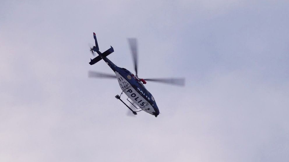 En blå och vit polishelikopter fotograferad underifrån mot en halvklar till mulen himmel.