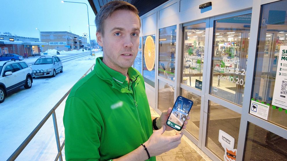 Butikschef Erik Norberg står utanför entrén till Coop i Norsjö. Han har en mobiltelefon i handen för att visa hur man loggar in med bank-id för att komma in i butiken. Det är vinter och i bakgrunden syns en snöig väg och två bilar.