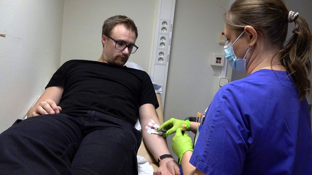 Bild på en man med svart t-shirt och svarta byxor som ligger på en sjukhussäng. Bredvid honom sitter en läkare med blå tröja och som håller i mannens omplåtstrade arm. Mannen heter Oliwer Andersson och är patient inom ett forskningsprojekt om diabetes.