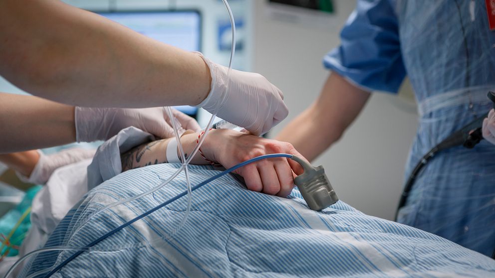 Bilden visar en sjukhussäng där man ser en hand på ett täcke, med en syremätare på fingret, samt två personer ur vårdpersonal som hjälper patienten. Danderyds sjukhus.