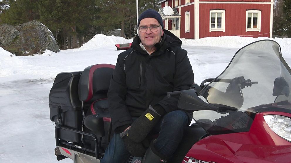 Johnny Stålarm bor på Hindersön i Luleå skärgård. Han sitter iklädd svart mössa och svarta vinterkläder på en djupröd skoter. I bakgrunden syns snö, barrskog och ett rött hus med vita knutar.