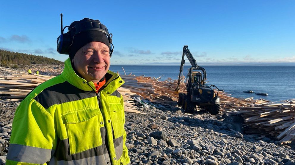 Mikael Danielsson som arbetar som bärgare står i arbetskläder ute vid kusten i Sjöviken utanför Härnösand. I bakgrunden syns en av maskinerna som plockar virket från vattnet och stranden. Virket ligger så långt ögat kan nå.