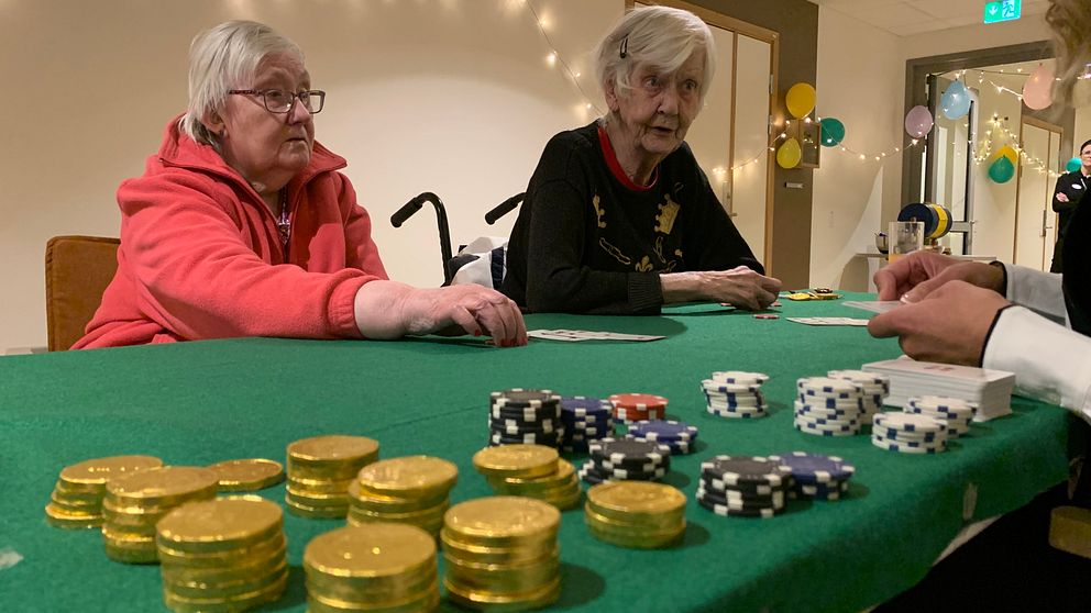 Två pensionärer på ett äldreboende sitter och spelar kort med spelmarker på det gröna spelbordet framför sig.