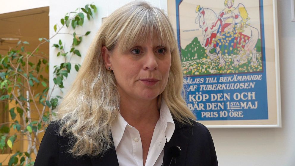 En kvinna (Majblommans generalsekreterare Åse Henell) Står i en korridor, bakom henne ginns en växt och en äldre affisch för majblomman, priset var 10 öre.