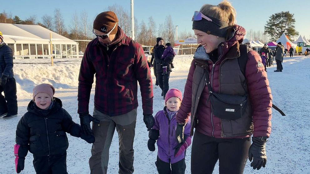 Två vuxna och två barn går hand i hand utomhus i vintrigt landskap. Bakom dem anas mer människor och mässtält.