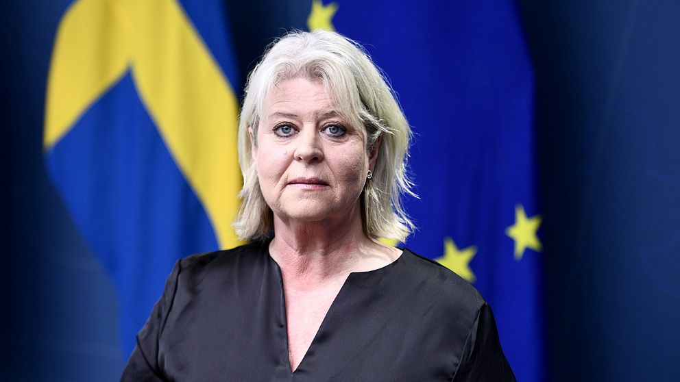 Socialtjänstminister Camilla Waltersson Grönvall (M) har ljust axellångt hår och svart glansig blus. Hon tittar in i kameran. Bakom henne syns en blå bakgrund med Sveriges och EU:s flaggor.