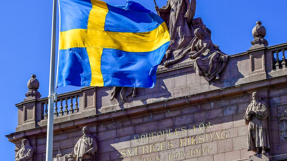 Svenska flaggan vajar framför en fasaddetalj på Riksdagen.