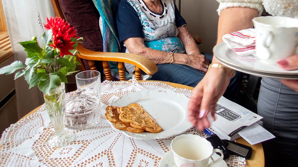 en äldre kvinna vid ett bord och ngn serverar henne kaffe.
