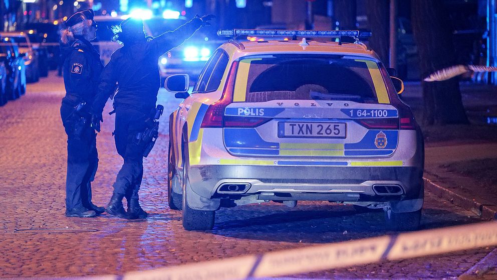 En polisbil står parkerad längs en trottoar, två kvinnliga poliser står i skenet från flera blå sirener och pekar inåt en gata.