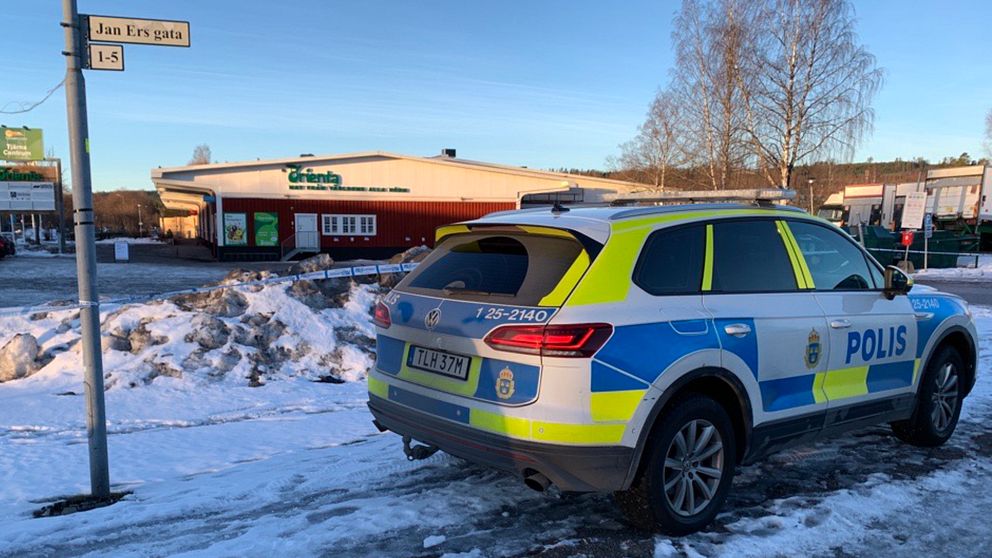 en polisbil som står parkerad, i bakgrunden syns avspärrningsband, och längre bort finns en matbutik. På en gatuskylt står det ”Jan Ers gata”.