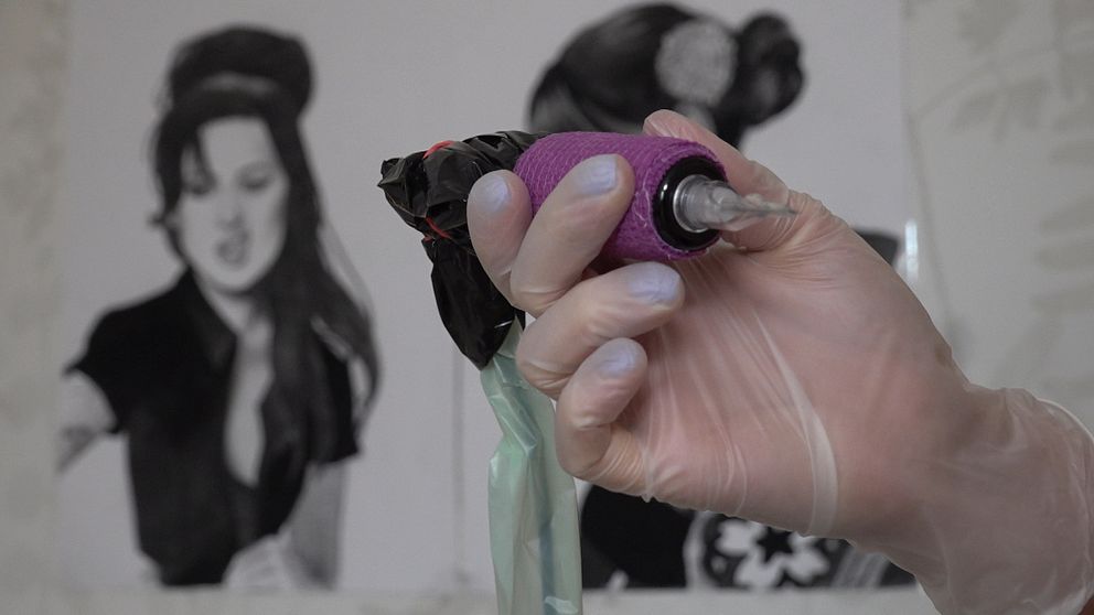 En handskbeklädd hand håller i en tatueringsmaskin med lila tjep. I bakgrunden syns en handmålad teckning på Amy Winehouse.