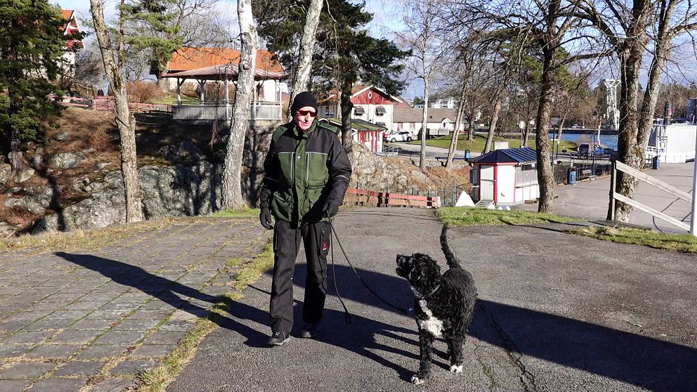 En äldre man med solglasögon och mössa går med en svart hund i koppel.