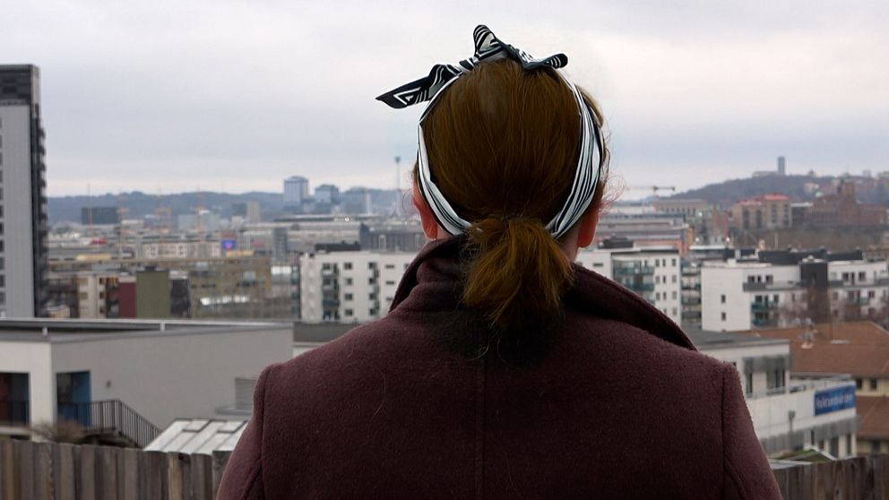 En kvinna står med ryggen mot kameran, hon har en kappa på sig och ett hårband knytet i håret. Hon tittar ut över Göteborg.
