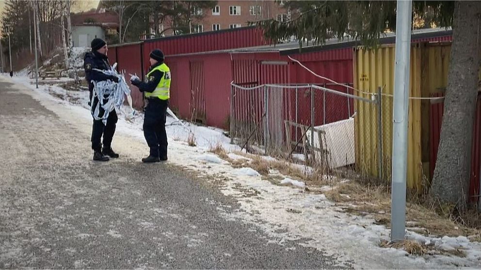 Två polisen står och samtalar på en liten grusväg. Den ene håller avspärrningsband i handen.