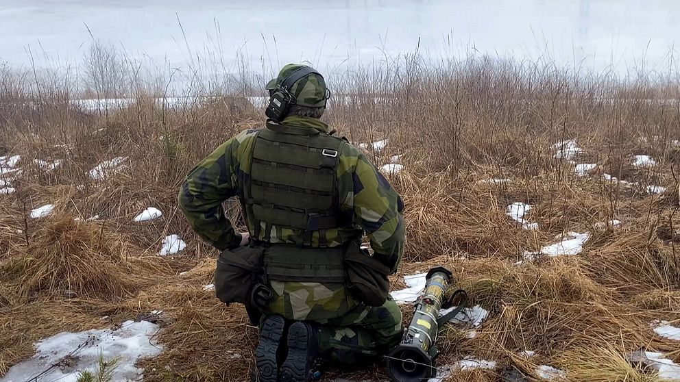 En soldat sitter på huk vid ett tomt fält med högt gräs och lite snö, med ryggen vänd mot kameran. På marken bredvid honom ligger en granatkastare.