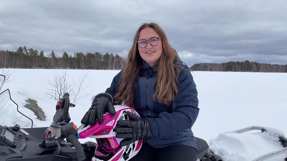 SVT Nyheter Västerbottens reporter Moa Lindberg, en ung kvinna med långt brunt hår och glasögon, sitter på en skoter på en istäckt sjö, iklädd vinterkläder, och håller en rosa skoterhjälm med händerna.