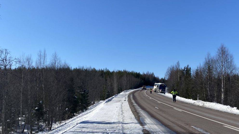 På bilden syns bussen i det högra hörnet, det är två personer som reflexvästar som går på vägen. Bilden är tagen utomhus och himlen är blå.