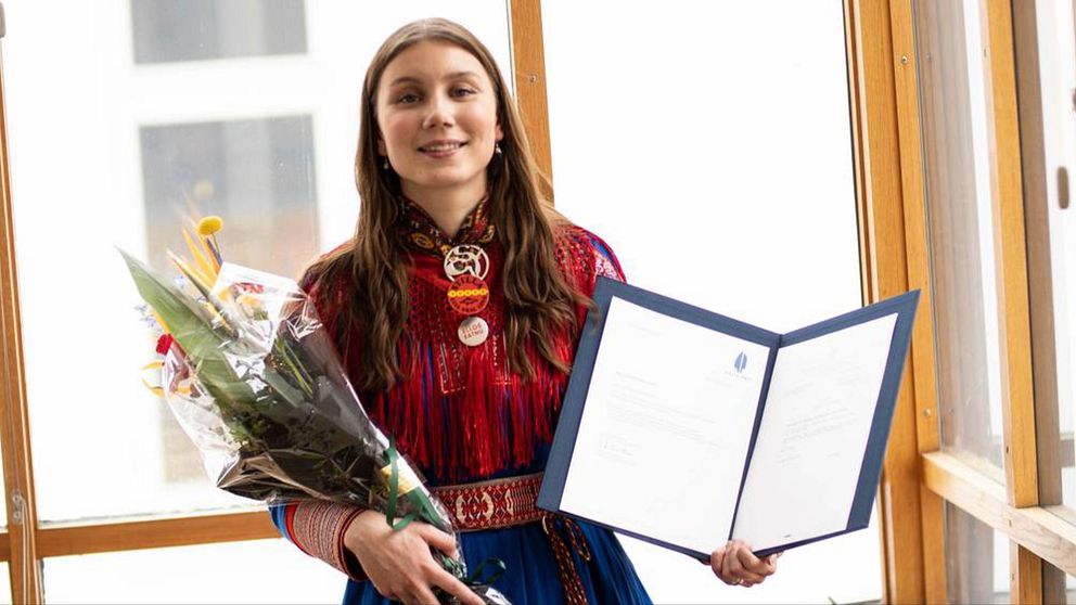 Ella Marie Hætta Isaksen som är en 25 årig tjej syns på bilden. Hon har långt brunt hår och bär på bilden en kolt från sitt samiska område. I ena handen håller hon en blombukett och i den andra en pärm med motiveringen till hennes vinst i den andra.