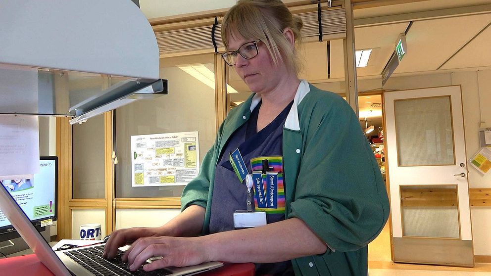 Kvinna med blont uppsatt hår och glasögon står vid en dator. Hon har grön rock på sig.