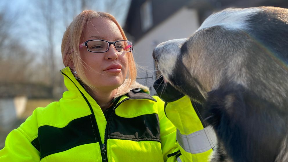 Bild på kvinna i 30-årsåldern med gul varseljacka. Hon är blond och har uppsatt hår. Hon har svarta och rosa glasögon. Hon håller upp sin vänstra hand och klappar på en hund.