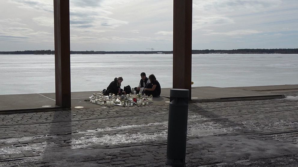 Tre personer sitter vid minnesplatsen i Södra hamn. Tända ljus står på marken.