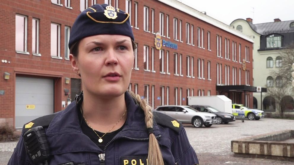 Kvinnlig polis framför Borlänge polishus. Hon har en polismössa med emblem på sig framför det tegelhuset.
