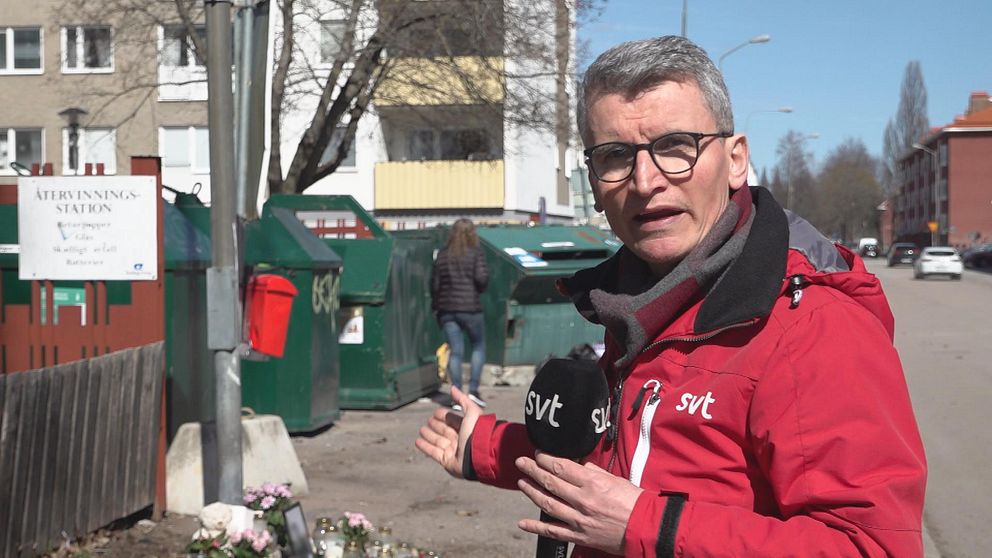 reporter framför återvinningsstation och blommor på marken
