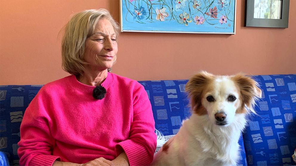 Birgitta Prytz sitter i en soffa framför en vägg med en hund till höger om henne som hon kollar ner på och ser glad ut.