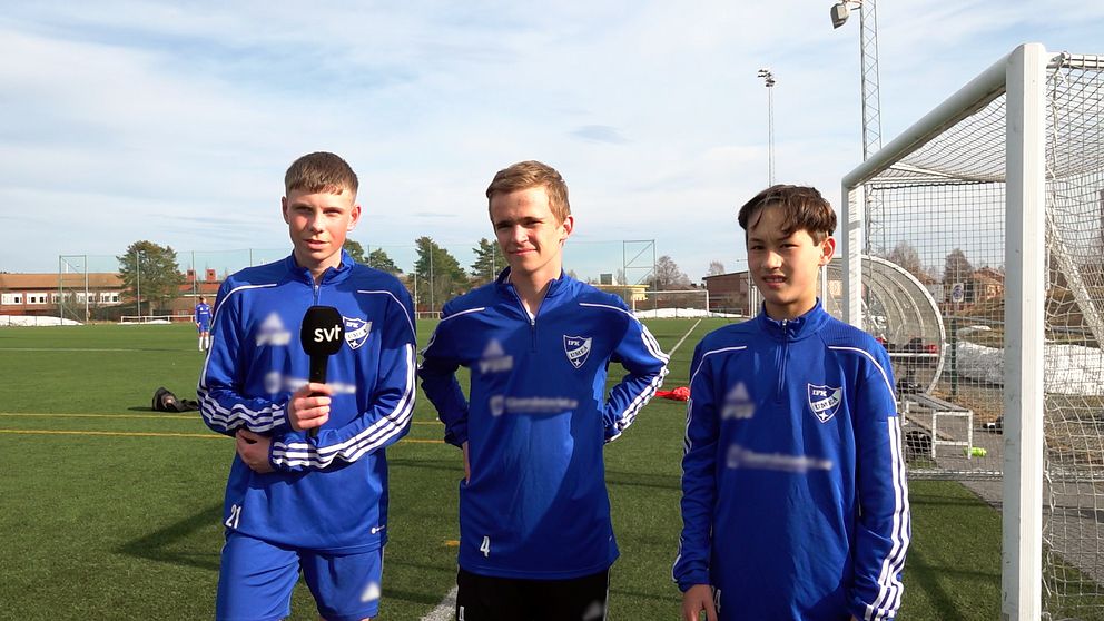 Tre unga killar i blåa fotbollsdräkter på en konstgräsplan