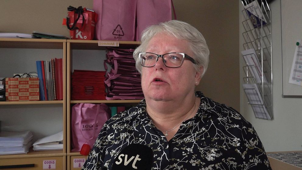 Carina Melander, en äldre kvinna med vitt hår och glasögon, intervjuas av SVT inne i ett rum med hyllor i bakgrunden.